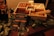 A wooden box contains separate compartments filled with marshmallows, graham crackers, chocolate, and a small can of fuel. Skewers are placed on top and a detailed guide for making s'mores is visible in front of the box. Green leaves are spread around the box for decoration.