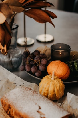 A decorative table setting featuring a variety of autumn-themed items, including two small pumpkins, a cluster of grapes, a small loaf of bread dusted with powdered sugar, and a vase with dried leaves. There are also candles and a black candle holder in the background.