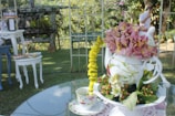 A decorative setup on a glass table featuring a teapot filled with pink and yellow flowers. A teacup with a matching floral design sits next to the teapot. Surrounding the main display are various pieces of white furniture, with a garden setting in the background.