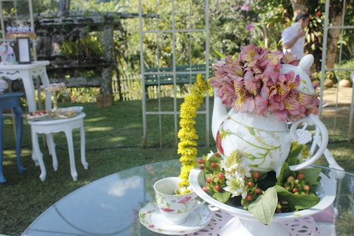 A decorative setup on a glass table featuring a teapot filled with pink and yellow flowers. A teacup with a matching floral design sits next to the teapot. Surrounding the main display are various pieces of white furniture, with a garden setting in the background.