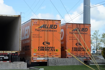Two orange freight trailers are parked closely together, branded with the 'Allied' logo and the words 'The Careful Movers'. There are wires overhead and a partially visible open trailer to the left. The ground is concrete, and there are some trees and buildings in the background.