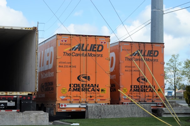 Two orange freight trailers are parked closely together, branded with the 'Allied' logo and the words 'The Careful Movers'. There are wires overhead and a partially visible open trailer to the left. The ground is concrete, and there are some trees and buildings in the background.