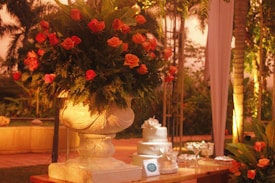 A large, ornate stone vase overflows with vibrant orange roses and lush greenery, placed on a wooden table. Beside it is a three-tiered white cake adorned with delicate floral decorations. The setting is an outdoor area with soft, warm lighting and foliage in the background, creating a serene and romantic atmosphere.