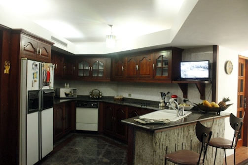 A modern kitchen with dark wooden cabinets and a corner layout. The counter features a built-in sink and a decorative fruit basket with bananas, pineapples, and other fruits. There is a flat-screen television mounted on the wall, a microwave, a refrigerator with photos and magnets on the door, and two high chairs at the kitchen island. The room is well-lit with a ceiling light.