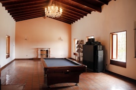 A spacious room with a high ceiling and exposed wooden beams features a pool table at its center. The floor is covered with terracotta tiles, and a chandelier hangs from the ceiling, adding an element of elegance. One side of the room includes windows with wooden frames allowing natural light to fill the space. There is a fireplace along one wall, and a set of shelves containing various items and a stereo system are positioned against another wall.