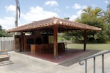 A covered outdoor seating area with a terracotta tiled roof and wooden support beams, featuring a brick oven or grill with a chimney. There are several wooden stools at the countertop, and the area is surrounded by a grassy yard and trees. The sky is clear with a few clouds.