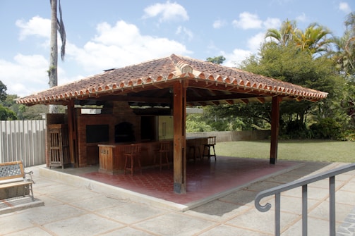 A covered outdoor seating area with a terracotta tiled roof and wooden support beams, featuring a brick oven or grill with a chimney. There are several wooden stools at the countertop, and the area is surrounded by a grassy yard and trees. The sky is clear with a few clouds.