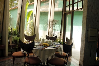A cozy dining area with a round table set for breakfast, featuring a fruit bowl with bananas and a papaya. The room is filled with natural light from large windows, surrounded by lush green plants. The chairs have a unique design with circular backrests, and the walls are painted in light green tones.
