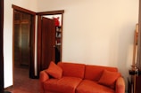 A room featuring an orange upholstered sofa with matching cushions, set against a plain white wall. There are wooden doors and a glimpse of wooden shelves in the background, along with some decorative items including a red stuffed toy. The decor is simple and the flooring is tiled.
