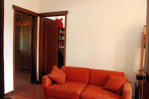 A room featuring an orange upholstered sofa with matching cushions, set against a plain white wall. There are wooden doors and a glimpse of wooden shelves in the background, along with some decorative items including a red stuffed toy. The decor is simple and the flooring is tiled.