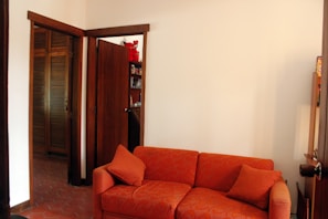 A room featuring an orange upholstered sofa with matching cushions, set against a plain white wall. There are wooden doors and a glimpse of wooden shelves in the background, along with some decorative items including a red stuffed toy. The decor is simple and the flooring is tiled.