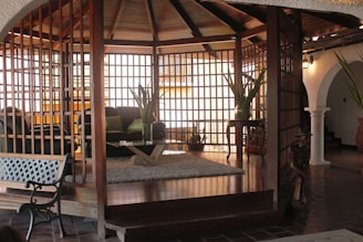 A living room area with a wooden lattice structure surrounds plush seating, including a black sofa adorned with green cushions. A glass coffee table with a wooden base sits on a soft, fluffy rug. Large potted plants add a touch of nature, and a wooden figure stands as a decorative piece. The space features warm, wooden tones contrasted by white archways and tiled flooring.