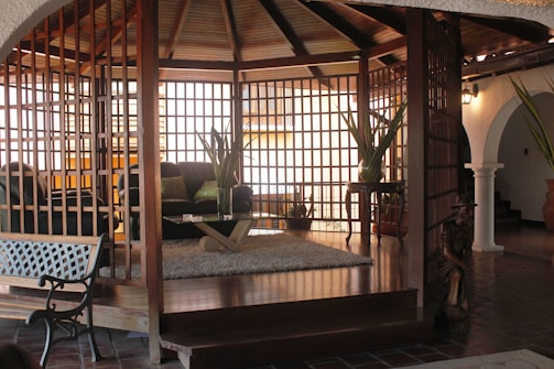 A living room area with a wooden lattice structure surrounds plush seating, including a black sofa adorned with green cushions. A glass coffee table with a wooden base sits on a soft, fluffy rug. Large potted plants add a touch of nature, and a wooden figure stands as a decorative piece. The space features warm, wooden tones contrasted by white archways and tiled flooring.