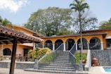 A large, Mediterranean-style villa with an orange facade and a clay-tiled roof. The house features several arched windows and a central staircase leading up to a wide patio. Potted plants and a variety of tropical trees surround the property, and large clay pots are placed near the staircase.