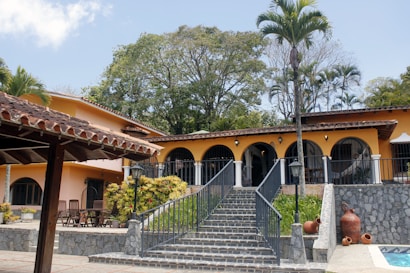 A large, Mediterranean-style villa with an orange facade and a clay-tiled roof. The house features several arched windows and a central staircase leading up to a wide patio. Potted plants and a variety of tropical trees surround the property, and large clay pots are placed near the staircase.