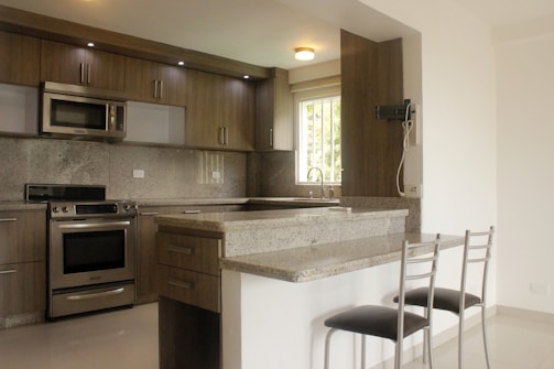 A modern kitchen with stainless steel appliances including a microwave and oven. The cabinetry is wooden with a grainy texture, and there is a granite countertop with an overhang for bar seating. Two bar stools with metallic legs and black seats are positioned at the counter. Natural light enters through a window above the sink, which has a metallic faucet.