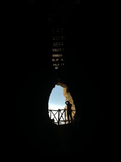 A silhouette of a person stands in front of a cave entrance, peering out towards a bright blue sky framed by rocky formations. A wooden railing is visible, adding a sense of structure and safety.