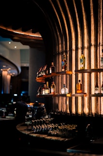 Shelves of a stylish bar display various bottles of liquor against an industrial-looking textured wall illuminated with warm, ambient lighting. Glasses are neatly arranged on the countertop below, adding to the classy and sophisticated atmosphere.