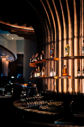 Shelves of a stylish bar display various bottles of liquor against an industrial-looking textured wall illuminated with warm, ambient lighting. Glasses are neatly arranged on the countertop below, adding to the classy and sophisticated atmosphere.