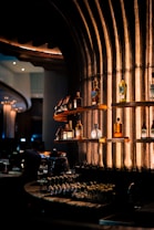 Shelves of a stylish bar display various bottles of liquor against an industrial-looking textured wall illuminated with warm, ambient lighting. Glasses are neatly arranged on the countertop below, adding to the classy and sophisticated atmosphere.