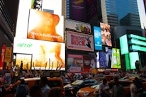 A bustling urban scene filled with a large number of bright and colorful billboards, displaying advertisements and promotions. The crowded street below is packed with people and yellow taxis, indicative of a lively city environment. Skyscrapers and modern buildings frame the backdrop, while digital screens show various ads.