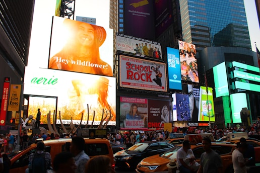 A bustling urban scene filled with a large number of bright and colorful billboards, displaying advertisements and promotions. The crowded street below is packed with people and yellow taxis, indicative of a lively city environment. Skyscrapers and modern buildings frame the backdrop, while digital screens show various ads.