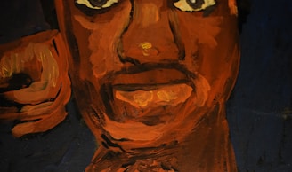 A close-up view of a painted face using bold strokes in shades of orange and brown on a dark background. The expression is intense with large, prominent eyes and lips slightly parted.