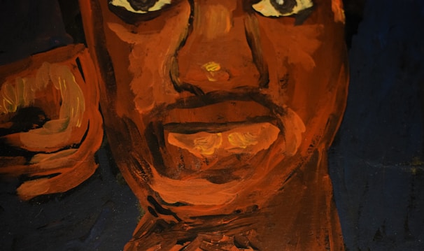 A close-up view of a painted face using bold strokes in shades of orange and brown on a dark background. The expression is intense with large, prominent eyes and lips slightly parted.