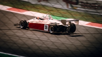 A formula race car with a red and white color scheme is speeding down a racetrack, with the number 50 prominently displayed on its side. The image is slightly blurred, emphasizing the speed and motion of the vehicle. The track has red and white painted curbs and a fence in the foreground.