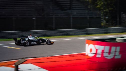 A race car speeds along a track, captured in motion. The car features prominent branding, and the blurred background suggests high speed. In the foreground, a red barrier with visible text provides contrast.