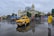 A vintage yellow and blue taxi is prominently visible on a wet, reflective street in front of a grand colonial-style building with a clock tower. Several other vehicles, including buses and motorcycles, are in the vicinity, and a man wearing a bright yellow shirt stands on the right side of the scene. The overcast sky suggests recent rain, adding to the reflective nature of the ground.