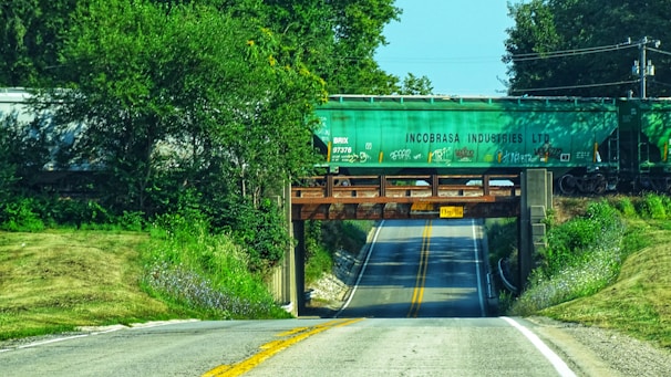 A rural road passing under a railway bridge carrying a green cargo train with the label 'Incobrasa Industries LTD.' The bridge is made of metal and concrete, and the surrounding vegetation is lush and green. The road has two lanes with yellow lines in the middle and leads up to the bridge with clear skies in the background.