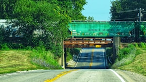 A rural road passing under a railway bridge carrying a green cargo train with the label 'Incobrasa Industries LTD.' The bridge is made of metal and concrete, and the surrounding vegetation is lush and green. The road has two lanes with yellow lines in the middle and leads up to the bridge with clear skies in the background.