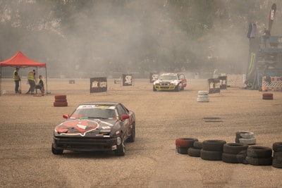 Several race cars are drifting on a concrete surface surrounded by stacks of tires and safety barriers. There is a red tent on the left side with people in reflective vests, and a lot of smoke or dust is in the air, partially obscuring the background.