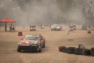 Several race cars are drifting on a concrete surface surrounded by stacks of tires and safety barriers. There is a red tent on the left side with people in reflective vests, and a lot of smoke or dust is in the air, partially obscuring the background.