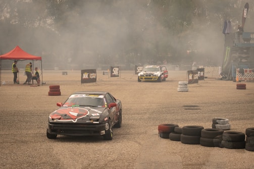 Several race cars are drifting on a concrete surface surrounded by stacks of tires and safety barriers. There is a red tent on the left side with people in reflective vests, and a lot of smoke or dust is in the air, partially obscuring the background.
