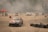 Several race cars are drifting on a concrete surface surrounded by stacks of tires and safety barriers. There is a red tent on the left side with people in reflective vests, and a lot of smoke or dust is in the air, partially obscuring the background.