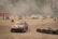Several race cars are drifting on a concrete surface surrounded by stacks of tires and safety barriers. There is a red tent on the left side with people in reflective vests, and a lot of smoke or dust is in the air, partially obscuring the background.
