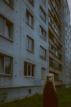 A young woman with long blonde hair stands in front of a multi-story residential building. The structure has a worn appearance with visible concrete and graffiti on the lower part. Several windows, some open and some closed, line the building's facade. The grass in the foreground is lush and green, contrasting with the muted tones of the building.