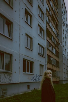 A young woman with long blonde hair stands in front of a multi-story residential building. The structure has a worn appearance with visible concrete and graffiti on the lower part. Several windows, some open and some closed, line the building's facade. The grass in the foreground is lush and green, contrasting with the muted tones of the building.
