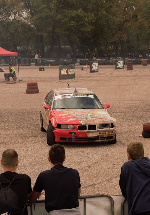 A red and white customized car is performing a drift maneuver on a gravel track, surrounded by barriers and signage for an automotive event. Spectators are watching the action from behind a fence. A red canopy and several barriers mark the area.