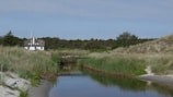 A serene landscape featuring a small house with a thatched roof nestled among tall trees and grass. In the foreground, a calm stream reflects the surrounding greenery, with sandy banks flanking its sides. The sky is clear, enhancing the peaceful and natural setting.