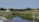 A serene landscape featuring a small house with a thatched roof nestled among tall trees and grass. In the foreground, a calm stream reflects the surrounding greenery, with sandy banks flanking its sides. The sky is clear, enhancing the peaceful and natural setting.