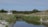 A serene landscape featuring a small house with a thatched roof nestled among tall trees and grass. In the foreground, a calm stream reflects the surrounding greenery, with sandy banks flanking its sides. The sky is clear, enhancing the peaceful and natural setting.