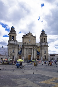 A grand, historic cathedral with two prominent towers stands under a partly cloudy sky. Numerous people are gathered around the area, engaging in various activities. Pigeons are scattered across the cobblestoned plaza. A few stalls with colorful umbrellas are visible, and the atmosphere is lively with a sense of social interaction.
