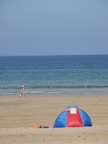 A peaceful beach scene features a small blue and red tent on the sandy shore. In the background, the calm ocean extends to the horizon under a clear blue sky. A person with a sunhat walks along the water's edge, accompanied by a child playing in the sand.