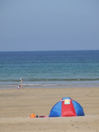 A peaceful beach scene features a small blue and red tent on the sandy shore. In the background, the calm ocean extends to the horizon under a clear blue sky. A person with a sunhat walks along the water's edge, accompanied by a child playing in the sand.