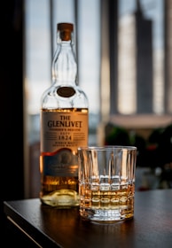 A bottle of Glenlivet whiskey and a glass with whiskey are placed on a wooden surface in a softly lit room. The background features tall windows with blurred cityscape views.