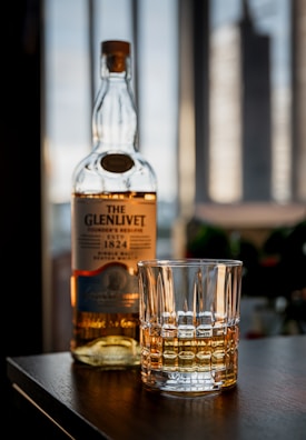 A bottle of Glenlivet whiskey and a glass with whiskey are placed on a wooden surface in a softly lit room. The background features tall windows with blurred cityscape views.