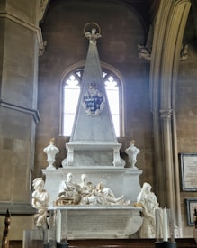 A detailed stone monument with classical sculptures is set against a Gothic-style window. It features reclining figures, ornate carvings, and a family crest. The monument is framed by arched architecture and flanked by carved urns.